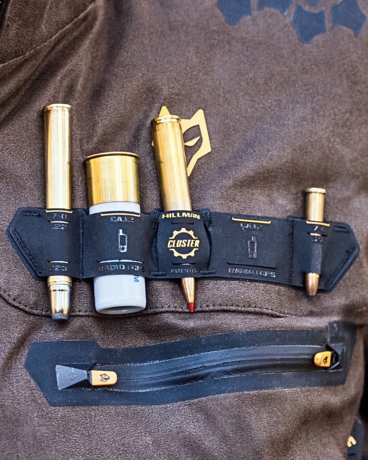 Braune Jacke mit einer schwarzen Tasche, die vier kugelförmige Gegenstände enthält und mit einer Reißverschlusstasche an der Brust befestigt ist. Die Tasche trägt die Aufschrift „HILLMAN CLUSTER“ und ist mit verschiedenen Symbolen und Patentinformationen versehen.