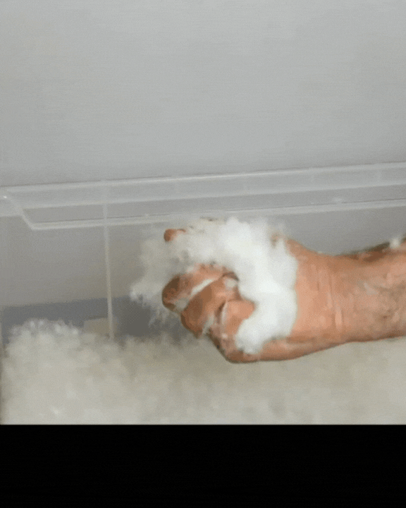 Hand, die eine Handvoll flauschiges weißes Material, das Baumwolle oder synthetische Füllung ähnelt, vor einem teilweise gefüllten Behälter hält.