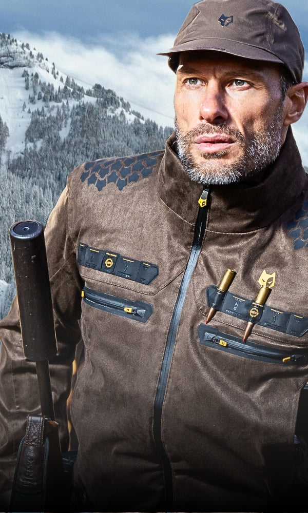 Ein Mann in brauner Outdoor-Ausrüstung und einer Mütze steht vor einer bergigen, schneebedeckten Landschaft im Hintergrund. Er hat einen Bart und hält ein Werkzeug oder einen Ausrüstungsgegenstand. Seine Jacke hat Reißverschlüsse und Taschen.