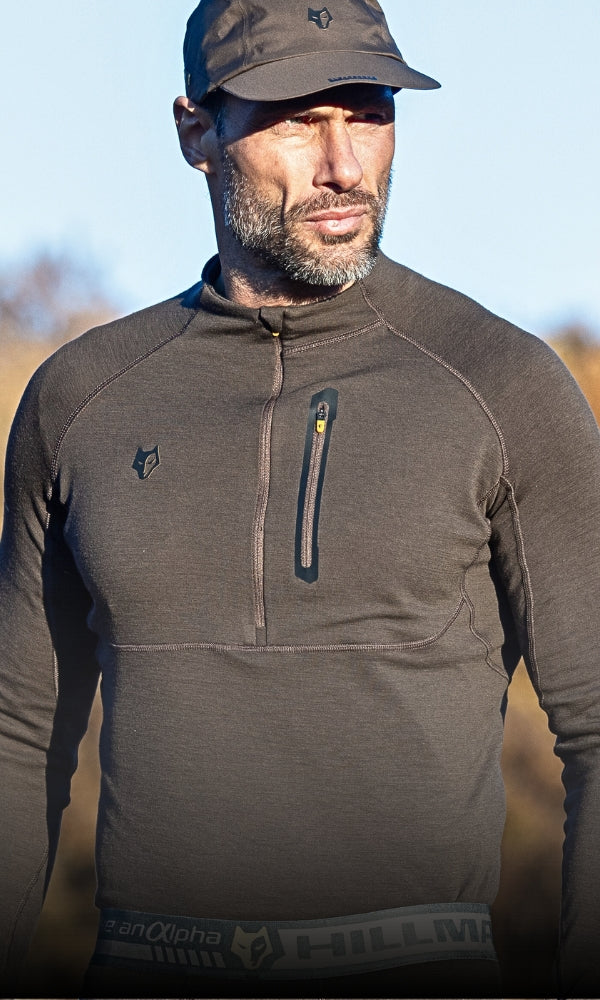Ein Mann mit einem braunen langärmligen Sportoberteil und einer Mütze steht im Freien bei natürlichem Licht.
