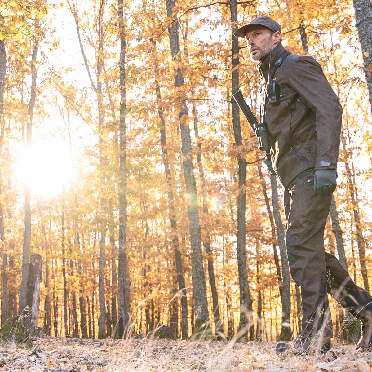 Eine Person in einer 6OL Winter-Jagdhose Camouflage von Hillman schlendert mit einer Mütze durch einen sonnendurchfluteten Herbstwald. Sie trägt eine Kamera und ein Stativ, umgeben von goldenen Blättern, durch deren Blätter das Sonnenlicht fällt.