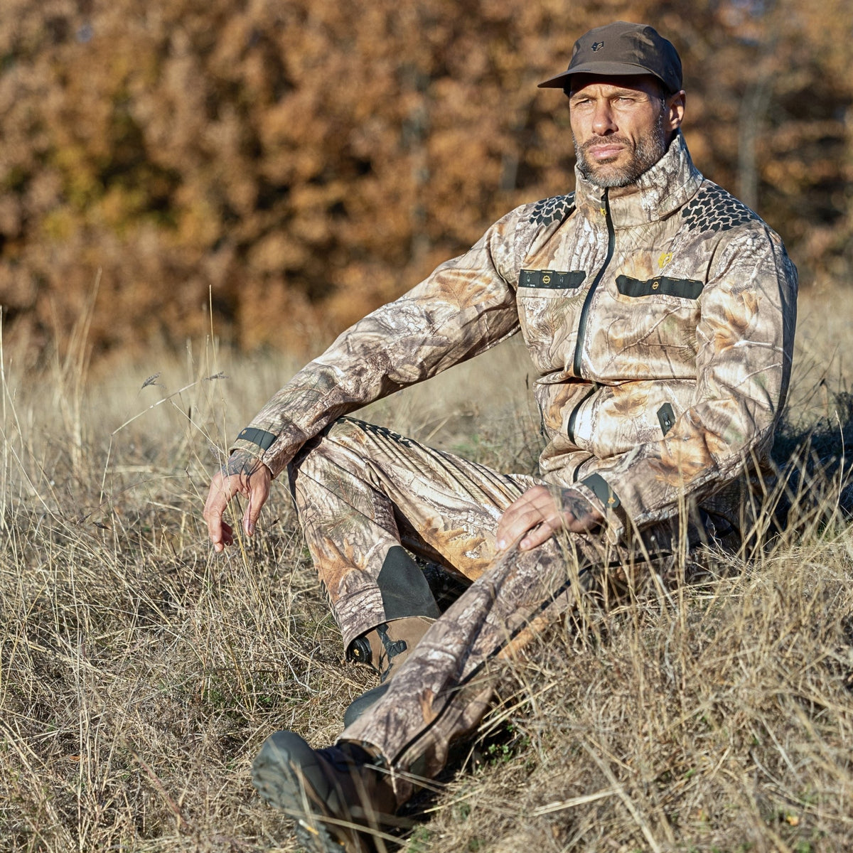 Ein Mann sitzt entspannt in seiner Tarnmütze und der Hillman 5WL Ultraleichte Regenjacke auf einer trockenen Wiese. Umgeben ist er von Bäumen mit leuchtendem Herbstlaub.