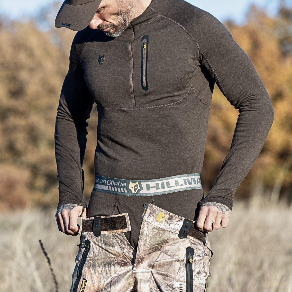 Ein Mann mit Mütze und dunklem Langarmhemd richtet seine Hillman-Tarnhose mit Hosenträgern. Diese Hose ist ideal für Outdoor-Abenteuer und passt perfekt zur 2BL Merino Lange Unterhose, einer Merino-Thermounterwäsche für die Jagd. Die Szene spielt vor hohem Gras und verschwommenen Bäumen.