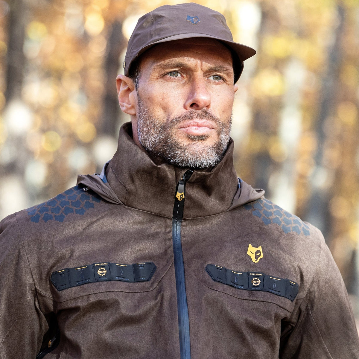 Ein Mann steht mit ernstem Gesichtsausdruck im Freien in einer braunen Hillman 5WL Ultraleichte Regenjacke und passender Mütze. Die Jacke hat schwarze Akzente und ein gelbes Logo. Verschwommene Herbstbäume im Hintergrund deuten auf einen klaren Tag hin, perfekt für Jagdabenteuer.