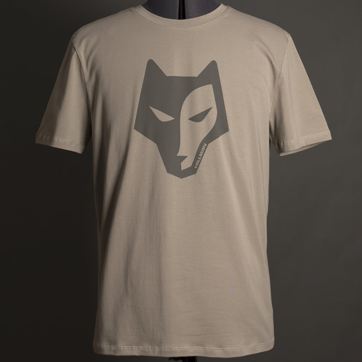 Das Hillman Jagdshirt Alpha – Jagd T-Shirt in Beige zeigt in der Mitte ein stilisiertes dunkles Wolfsgesicht. Es ist aus ägyptischer Baumwolle gefertigt und auf einer Schaufensterpuppe mit dunkelgrünem Hintergrund abgebildet, was die Hightech-Produktionsqualität unterstreicht.