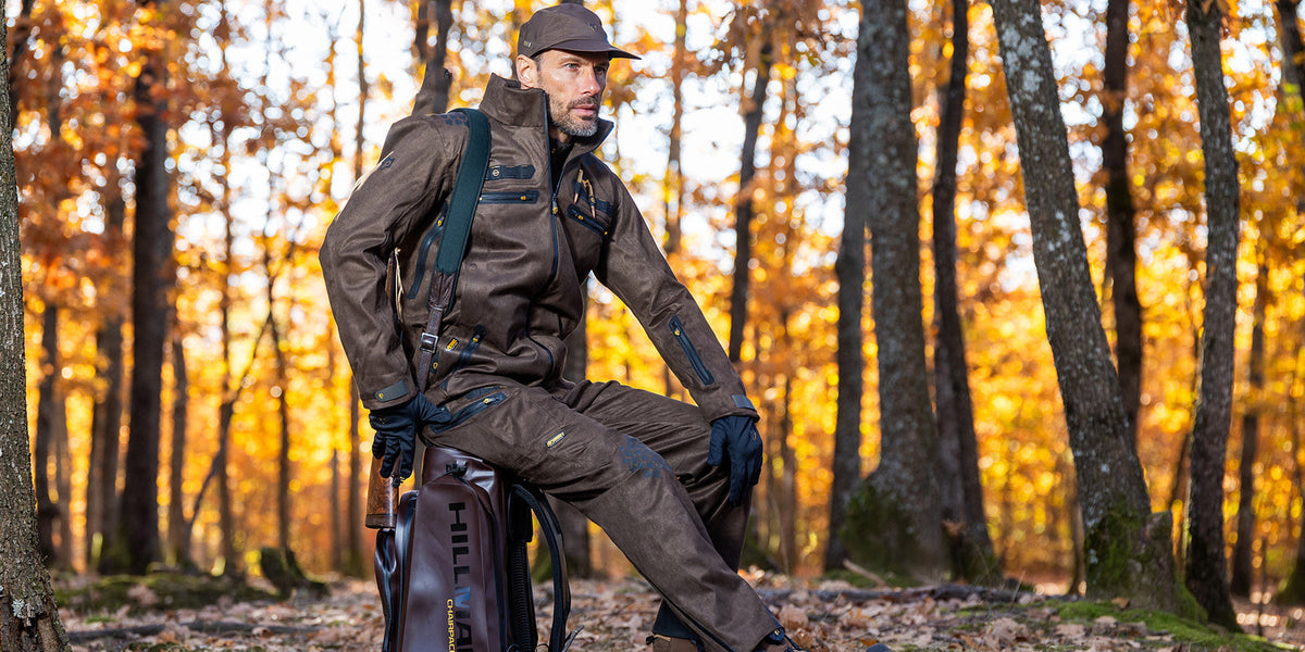 Jäger in funktionaler Jagdbekleidung im herbstlichen Wald sitzend auf Gepäckkoffer, passende Jagdausrüstung und Kleidung für unterschiedliche Klimabedingungen und Outdoor-Jagd.