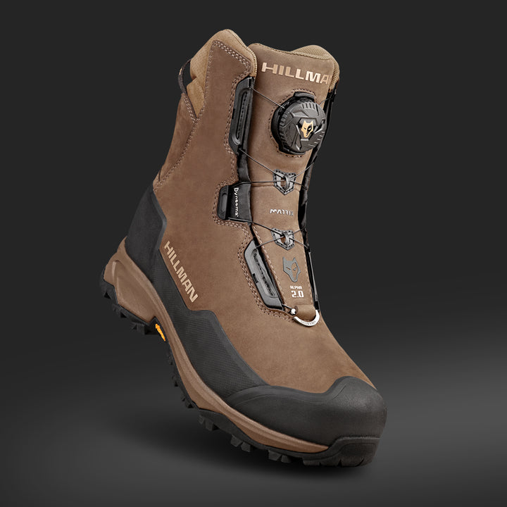 Ein brauner Wanderschuh mit schwarzen Akzenten und robuster Sohle, mit Schnürung und Drehverschluss. Der Markenname „HILLMAN“ ist an der Seite zu sehen, mit Logos auf der Zunge und in Knöchelnähe. Auf einem dunkelgrauen Hintergrund.