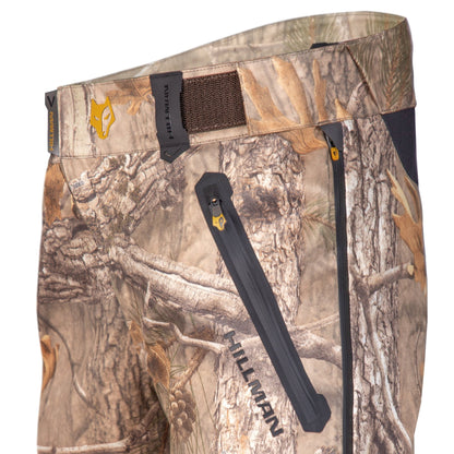 Nahaufnahme der wasserdichten Regenhose 5WL von Hillman – Jagdhose. Diese Tarnhose für die Jagd verfügt über eine Reißverschlusstasche und einen Klettverschluss am Bund. Das Waldmotiv ist auf dem Logo und dem Etikett mit dem Markennamen „Hillman“ zu sehen.