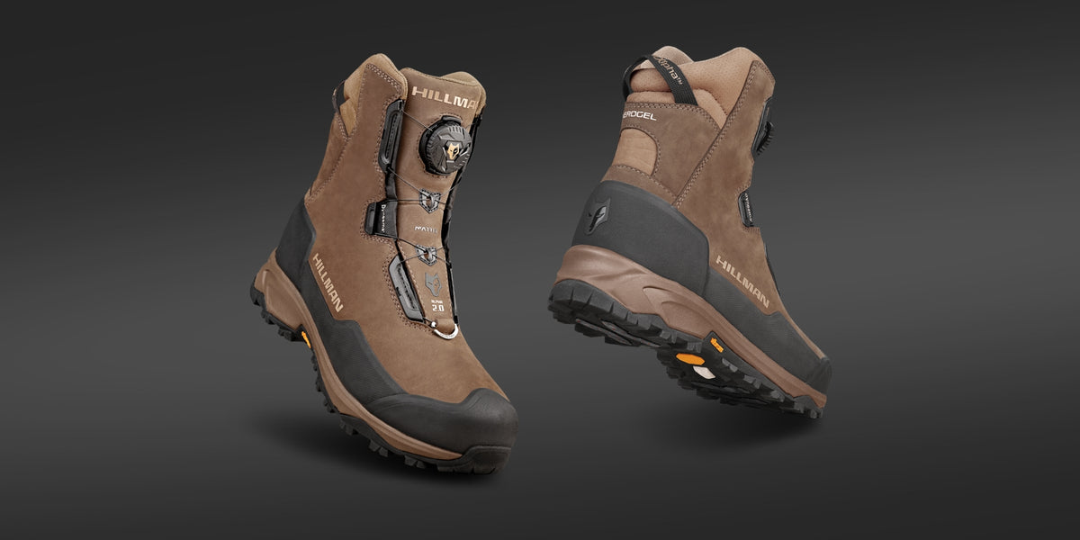 Ein Paar braune Wanderstiefel vor einem dunkelgrauen Hintergrund. Die Stiefel haben einen hohen Schaft, schwarze, verstärkte Zehenkappen und detaillierte Nähte. Ein Stiefel ist nach vorne gerichtet, der andere zeigt Ferse und Sohle.