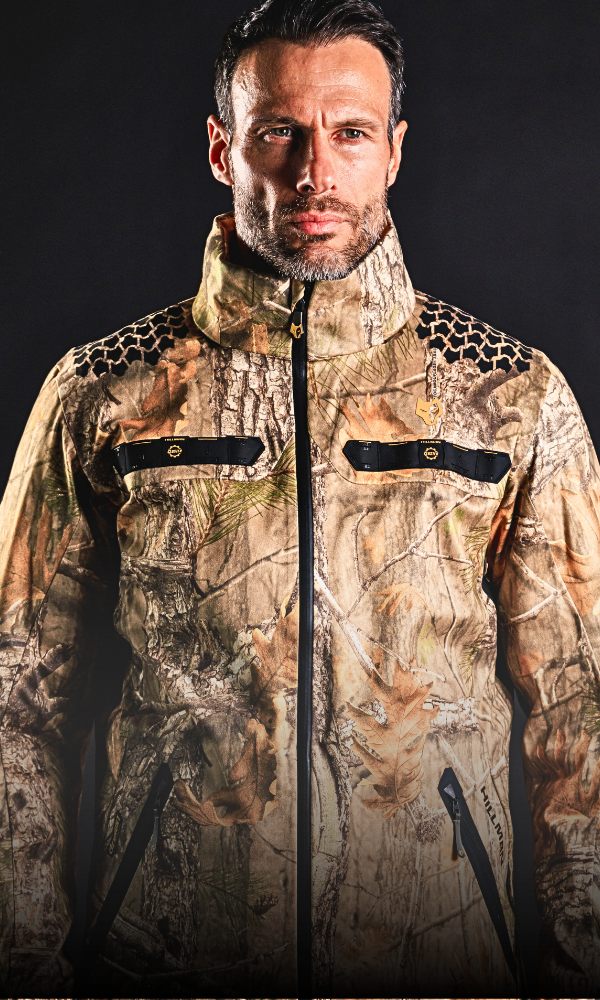 Eine Person in einer Tarnjacke steht vor einem dunklen Hintergrund. Die Jacke hat Reißverschlusstaschen und schwarze Details an den Schultern. Die Person hat kurzes dunkles Haar und einen konzentrierten Gesichtsausdruck.