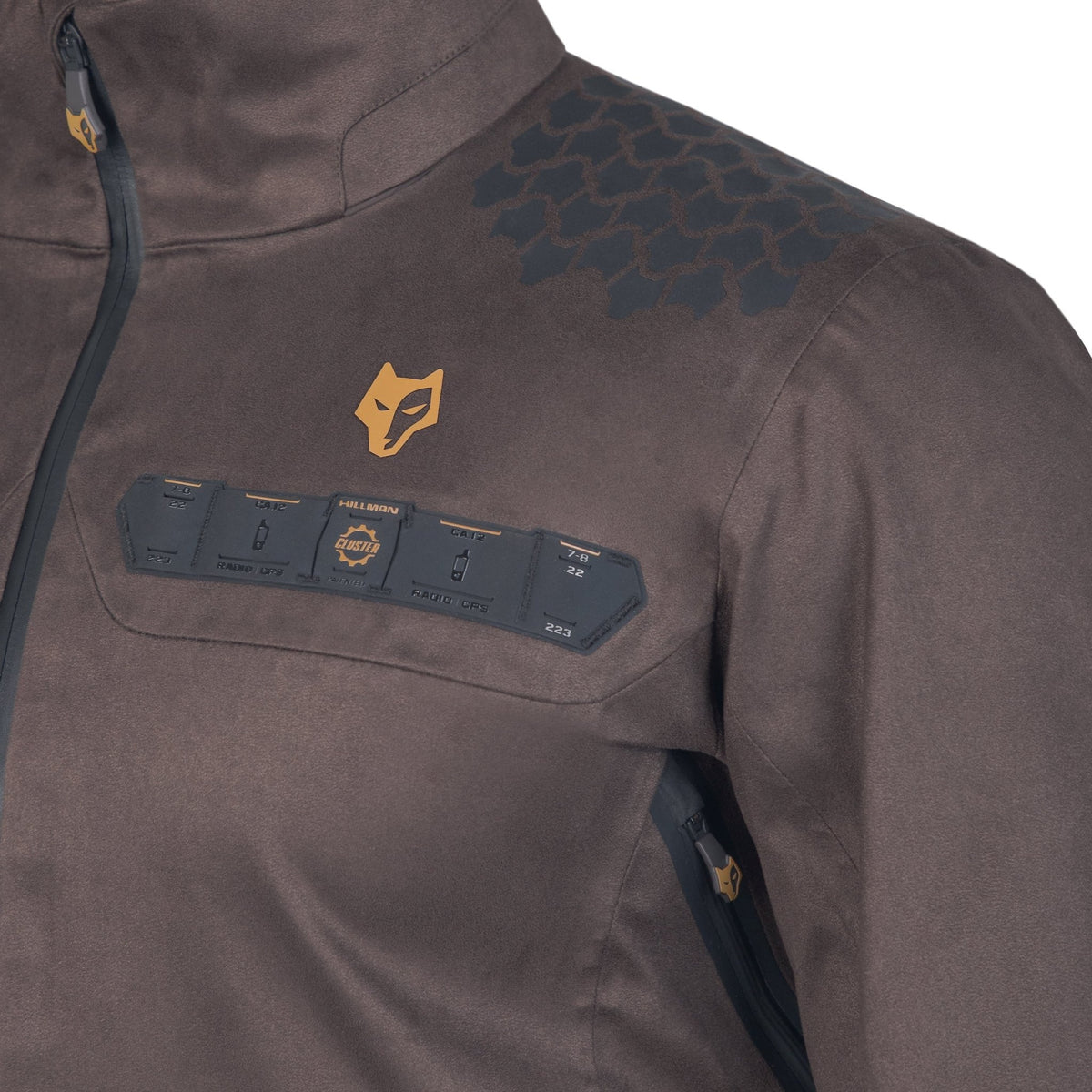 Nahaufnahme einer braunen Hillman 5WL Ultraleichte Regenjacke mit sechseckigem Muster auf der Schulter, einem Fuchs-Logo und einem Feld mit verschiedenen kleinen Symbolen und Text. Auf der linken Seite des Bildes befindet sich ein teilweise sichtbarer Reißverschluss.