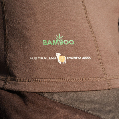 Nahaufnahme eines braunen Hillman Merino Oberteils mit grünem Text „BAMBOO“ und weißem Text „AUSTRALIAN MERINO WOOL“ und einem Schaf-Logo. Die Stoffmischung im 2BL Merino Langarmshirt bietet ein effektives Wärmeregulierungssystem.