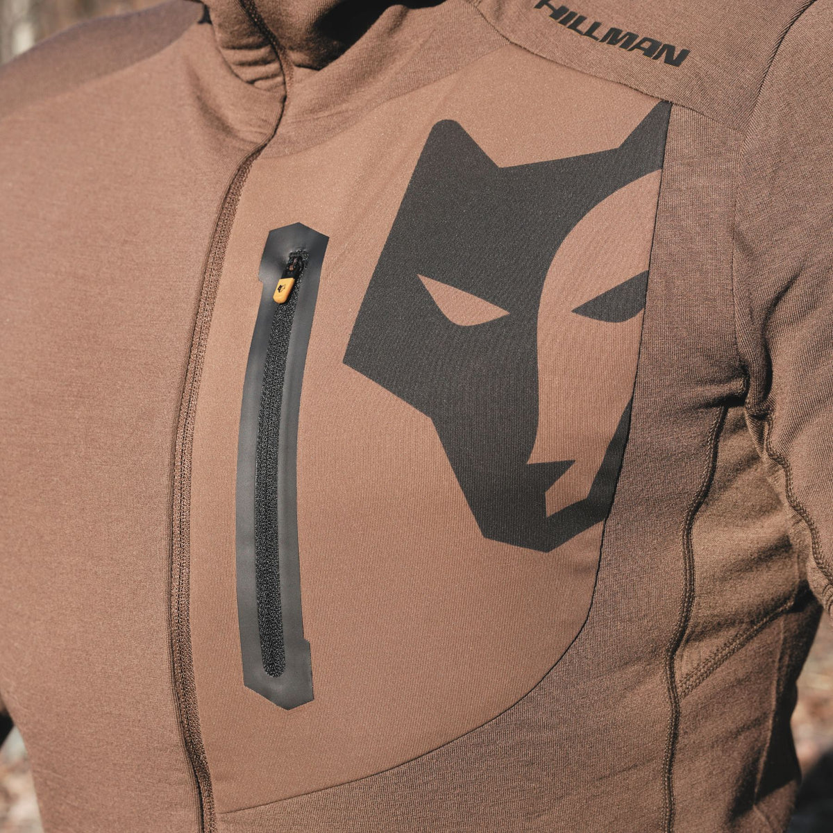 Eine Nahaufnahme zeigt jemanden, der einen braunen Hillman 3ML Jagd Hoodie mit Wolfskopf-Motiv auf der Brust trägt, hergestellt mit SYNERTECH™-Technologie. Der Hoodie verfügt über eine vertikale Reißverschlusstasche, alles vor einer verschwommenen Naturkulisse.