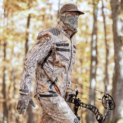 Eine vollständig getarnte Person mit passender Maske und Handschuhen trägt Hillmans 6OL Winter-Jagdhose Camouflage und hält einen Compoundbogen in einem Herbstwald, wobei sie nahtlos mit der Umgebung verschmilzt.