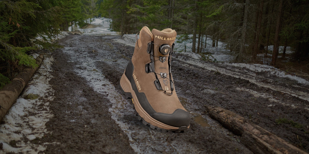 Ein brauner Wanderschuh sticht deutlich auf einem schlammigen Waldweg hervor. Die umliegenden Bäume sind mit Schnee bedeckt, was der rauen Naturlandschaft eine winterliche Kulisse verleiht. Der Schuh hat schwarze Schnürsenkel und eine robuste Sohle, was seine Strapazierfähigkeit unterstreicht.