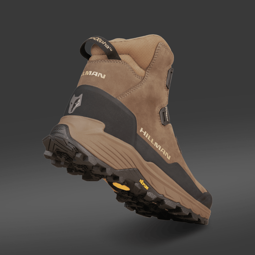 Ein brauner und schwarzer Jagdstiefel Wasserdicht - Herren Jagdschuhe | Alpha DryHunt™ Wanderstiefel von Hillman wird aus einem niedrigen Winkel von hinten auf einem grauen Hintergrund gezeigt, mit einer robusten Sohle und einem Wolfslogo an der Ferse.