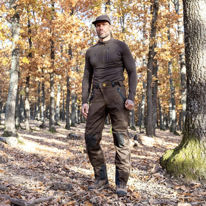 In einem sonnenbeschienenen Wald mit hohen Bäumen und gefallenen Blättern steht eine Person selbstbewusst mit Mütze, Langarmshirt und Hillmans 5WL Wasserdichte Regenhose – Jagdhose Dornenfest und Wasserdicht. Herbstblätter bilden eine warme Kulisse, während Licht durch die Zweige fällt.