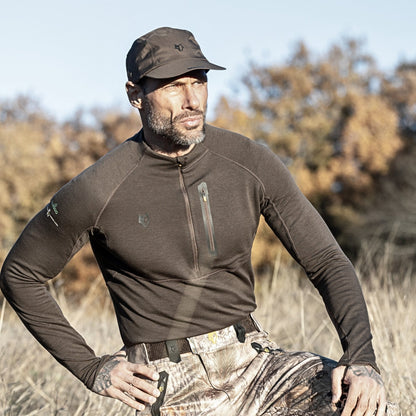 Ein Mann in Outdoor-Ausrüstung, darunter ein Hillman 2BL Merino Langarmshirt – Thermo-Unterwäsche, steht mit dunkler Mütze und Camouflage-Hose auf einer Wiese. Vor dem Hintergrund der verschwommenen Herbstbäume ist sein Gesichtsausdruck ernst.