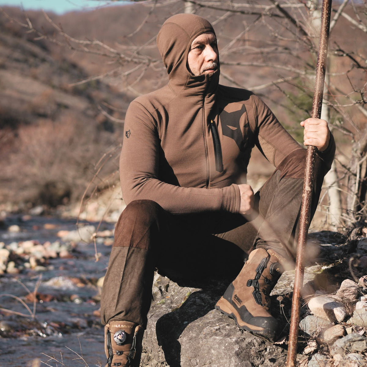 Eine Person in einem Hillman 3ML Jagd Hoodie mit SYNERTECH™-Technologie, möglicherweise aus Merinowolle, sitzt nachdenklich auf einem Felsen neben einem Bach und hält einen Wanderstock in der Hand. Die raue Landschaft ist geprägt von kahlen Bäumen, und der Wanderer scheint für kühles Wetter gekleidet zu sein.