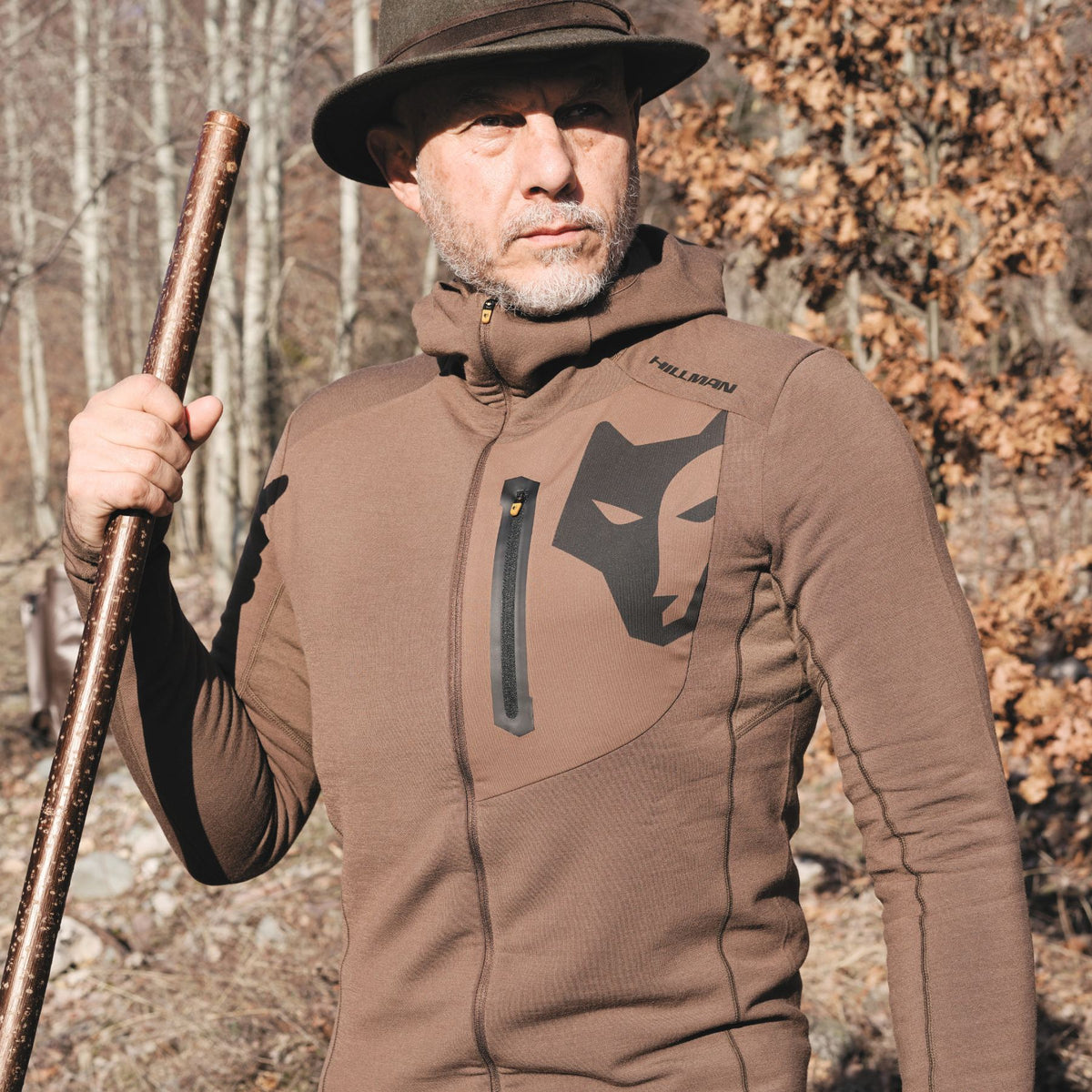 Ein Mann in einem Hillman 3ML Jagd-Hoodie mit Wolfslogo steht im Freien und hält einen Spazierstock in der Hand. Er trägt eine Mütze aus Merinowolle und ist von kahlen Bäumen und braunem Laub umgeben – eine herbstliche Kulisse, perfekt zum Testen der SYNERTECH™-Technologie.