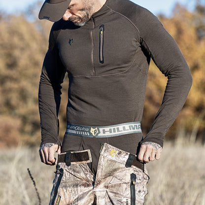 Ein Mann mit Mütze und dunklem Langarmhemd richtet seine Hillman-Tarnhose mit Hosenträgern. Diese Hose ist ideal für Outdoor-Abenteuer und passt perfekt zur 2BL Merino Lange Unterhose, einer Merino-Thermounterwäsche für die Jagd. Die Szene spielt vor hohem Gras und verschwommenen Bäumen.