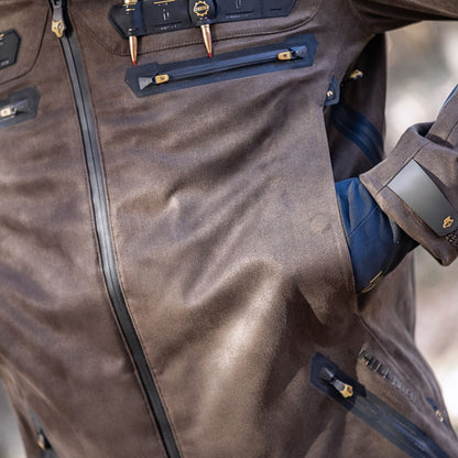 Eine Nahaufnahme zeigt jemanden in einer robusten Hillman 6OL Winter-Jagdjacke in Camouflage – Jagd-Tarnjacke mit schwarzen Reißverschlüssen und mehreren Taschen. Metallische Akzente zieren die Jacke, und in einer Tasche steckt eine Hand in einem blauen Handschuh.