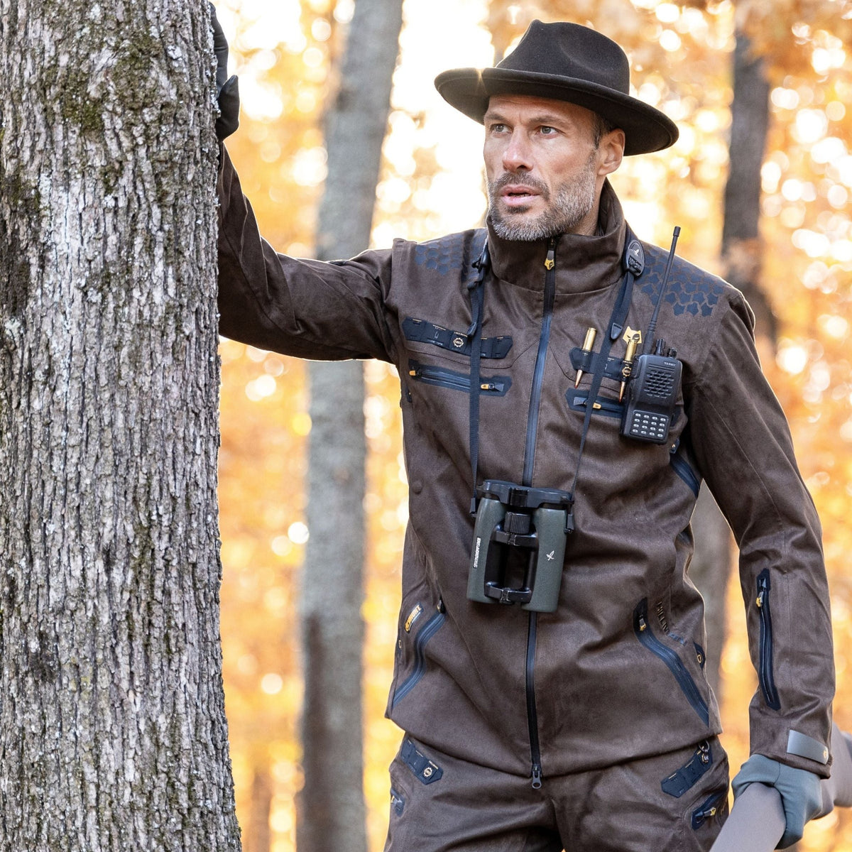 Eine Person in einer Hillman 6OL Winterjagdjacke in Camouflage und einem Hut hält ein Fernglas neben einem Baum. Ein Funkgerät ist an der Jacke befestigt und fängt die Atmosphäre eines herbstlichen Jagderlebnisses ein, umgeben von orangefarbenen und gelben Blättern.