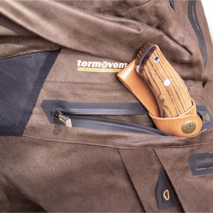 Nahaufnahme einer Person in wetterfester Kleidung mit dem „Termovent“-Logo. Ein Messer mit Holzgriff in einer hellbraunen Lederscheide steckt in einer Reißverschlusstasche ihrer Hillman 6OL Winter Jagdhose Camouflage – Tarnhose, wasserdichte Jagdhose.