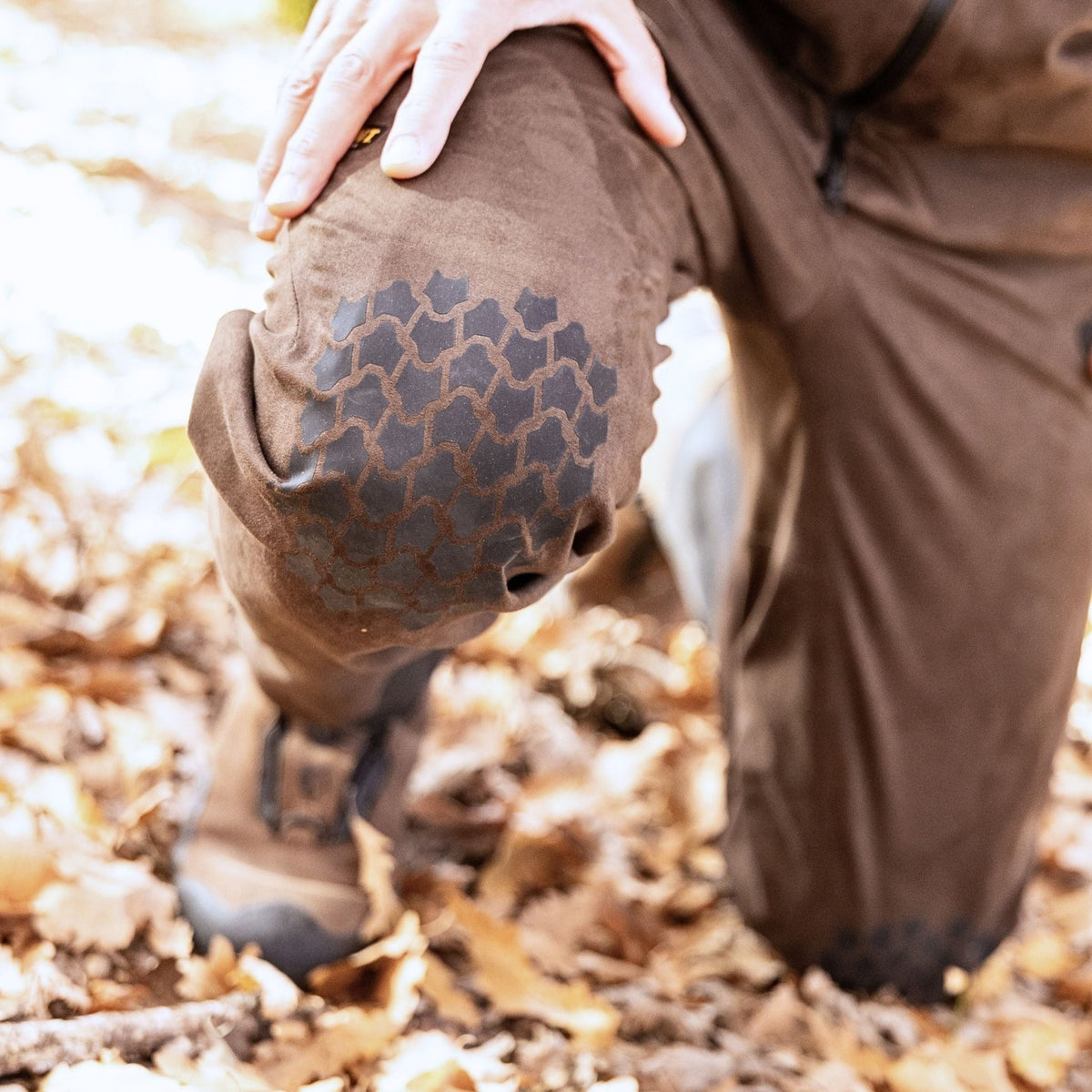 Eine Person kniet auf einem belaubten Boden. Sie trägt braune Hosen mit schwarzen Flicken im Wabenmuster an den Knien. Ihre linke Hand ruht auf ihrer Hillman 6OL Winter Jagdhose Camouflage. Sie trägt braune Stiefel, während Sonnenlicht durch die Blätter fällt.