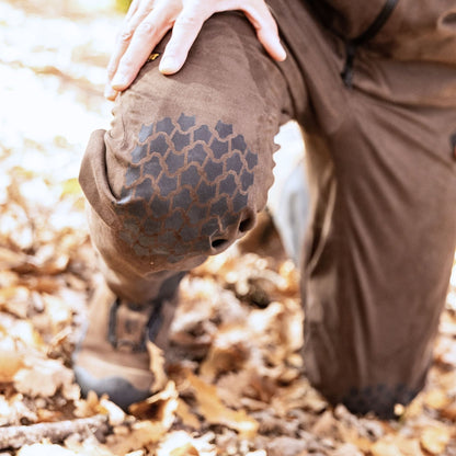 Eine Person kniet auf einem belaubten Boden. Sie trägt braune Hosen mit schwarzen Flicken im Wabenmuster an den Knien. Ihre linke Hand ruht auf ihrer Hillman 6OL Winter Jagdhose Camouflage. Sie trägt braune Stiefel, während Sonnenlicht durch die Blätter fällt.