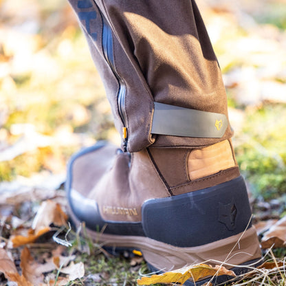 Eine Nahaufnahme zeigt eine Person in Hillmans braunen Wanderstiefeln mit Fuchskopf-Logo, umgeben von Laub. Die 6OL Winter Jagdhose Camouflage – Tarnhose von Hillman verfügt über einen teilweisen Reißverschluss, der auf wasserdichte oder isolierende Eigenschaften auf dem herbstlichen Grasboden hindeutet.