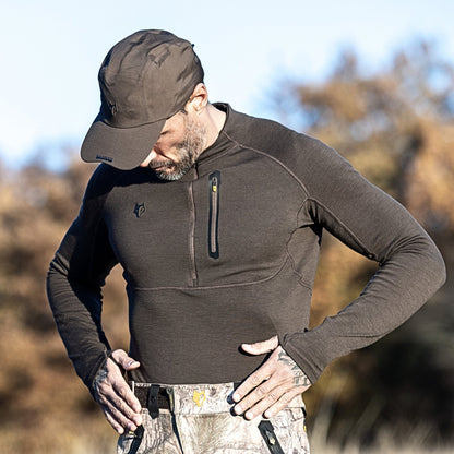 Eine Person in einem dunklen Hillman 2BL Merino Langarmshirt und einer Mütze blickt mit der Hand am Gürtel nach draußen. Die verschwommenen Bäume und der klare Himmel unterstreichen das Wärmeregulierungssystem des Shirts für Komfort bei wechselnden Temperaturen.