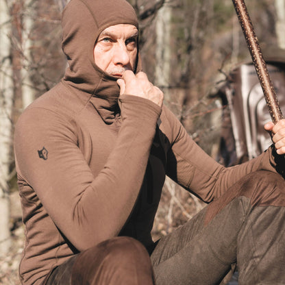 Eine Person in einem Hillman 3ML Jagd Hoodie aus Merinowolle sitzt in einem Waldstück und beißt sich auf den Daumen. Sie hält einen Holzstock in der Hand. Sonnenlicht wirft Schatten auf ihr Gesicht und hebt das zerklüftete Gelände um sie herum hervor.