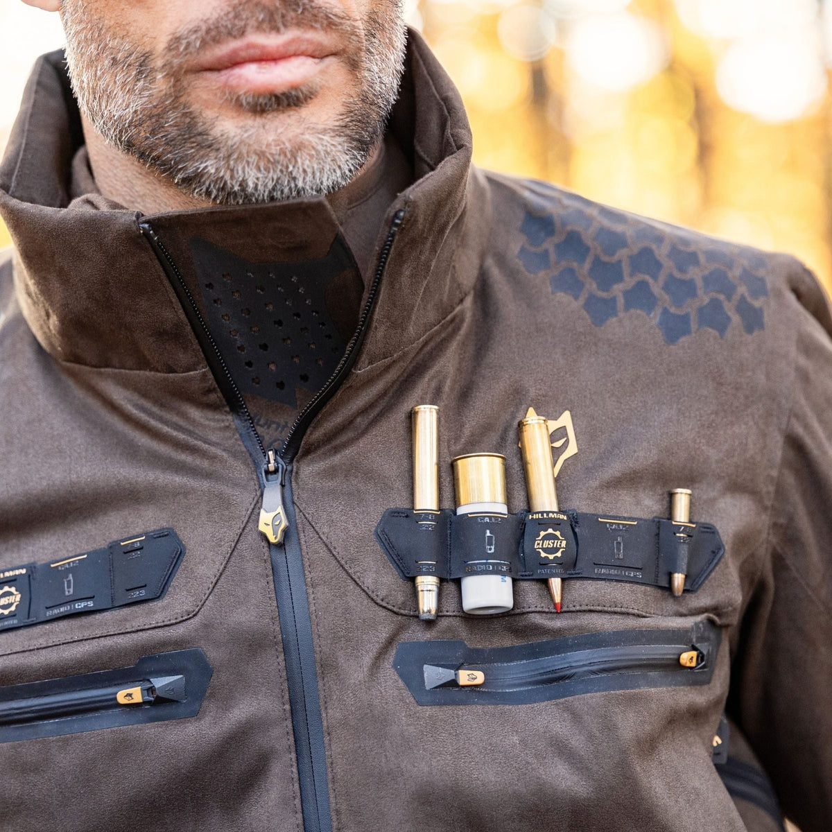 Eine Person trägt eine Hillman 6OL Winter-Jagdjacke in Camouflage, eine Jagdtarnjacke mit Mehrzweck-Werkzeugtaschen für Outdoor-Gadgets wie Stifte, eine Flasche und einen Brustgurt. Der sanft verschwommene Hintergrund lässt auf ein abenteuerliches Jagderlebnis in der Wildnis schließen.