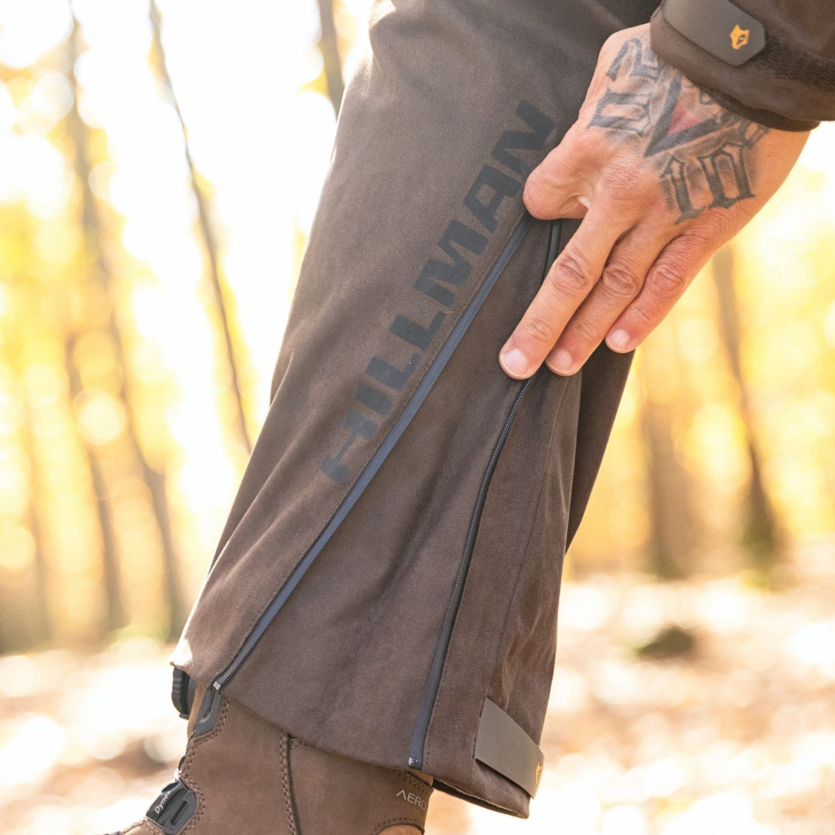 Im Wald, wo das Sonnenlicht durch die Bäume fällt, passt eine tätowierte Person den Reißverschluss ihrer Hillman 6OL Winter Jagdhose Camouflage – Tarnhose, wasserdichte Jagdhose an, während sie robuste braune Stiefel trägt.