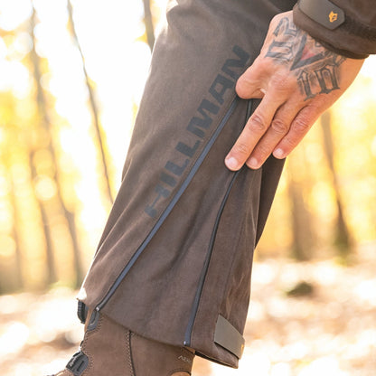 Im Wald, wo das Sonnenlicht durch die Bäume fällt, passt eine tätowierte Person den Reißverschluss ihrer Hillman 6OL Winter Jagdhose Camouflage – Tarnhose, wasserdichte Jagdhose an, während sie robuste braune Stiefel trägt.