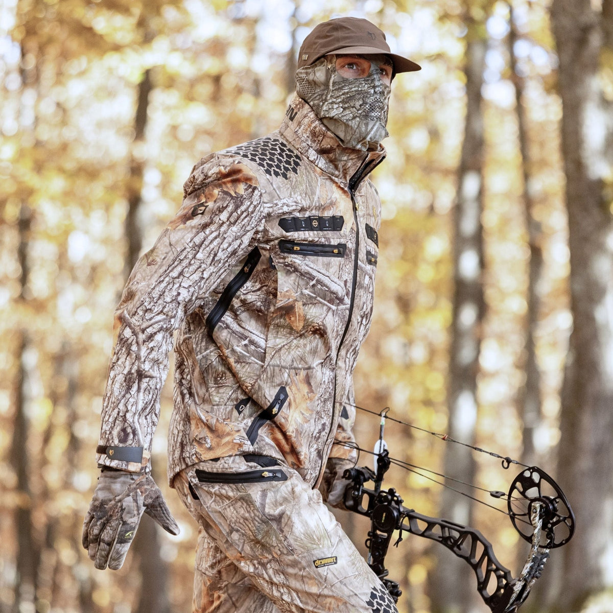 Sie tragen die 6OL Winter-Jagdjacke in Camouflage von Hillman, zusammen mit einer Maske, Handschuhen und einer Mütze, halten einen Compoundbogen und verschmelzen nahtlos mit dem herbstlich gefärbten Wald für ein authentisches Jagderlebnis.
