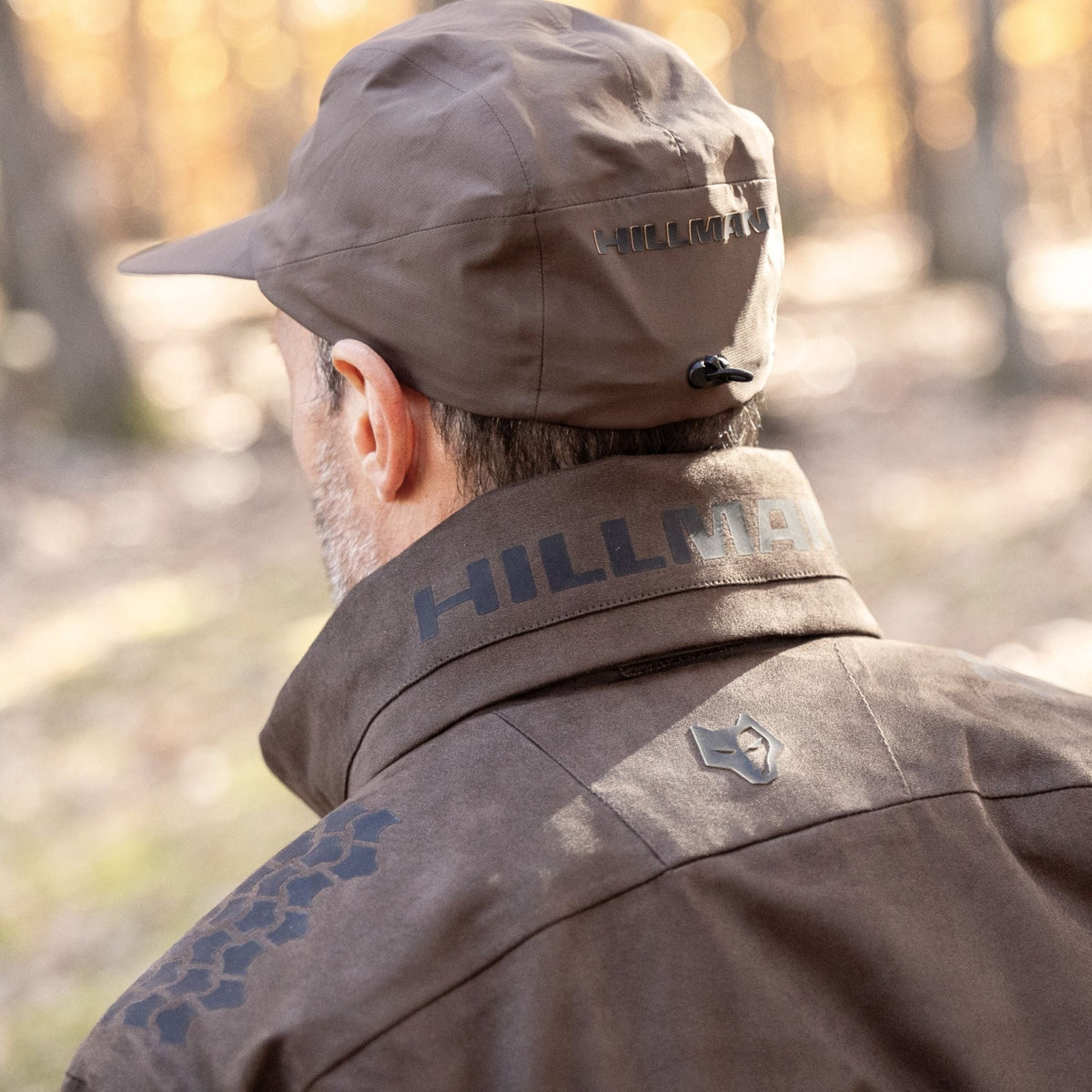 Von hinten ist eine Person zu sehen, die eine braune Mütze und die Hillman 6OL Winterjagdjacke in Camouflage trägt und mit dem herbstlichen Laub eines Waldhintergrunds verschmilzt.