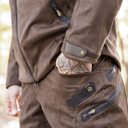 Eine Nahaufnahme zeigt jemanden in einer braunen Jacke und einer 6OL Winter Jagdhose Camouflage von Hillman, wobei die schwarzen Reißverschlussdetails hervorgehoben werden. Die Hand ruht in der Tasche und gibt den Blick auf ein Tattoo vor einem unscharfen Außenhintergrund frei.
