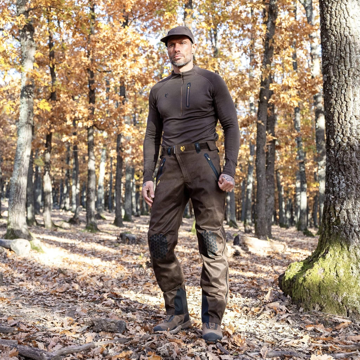 Die Person steht selbstbewusst in einem Wald mit herbstlichen Blättern unter den Füßen. Sie trägt eine Cargohose und ein braunes 2BL Merino Langarmshirt von Hillman. Sonnenlicht fällt durch die Bäume und sorgt dank des Wärmeregulierungssystems des Shirts für wohligen Tragekomfort.