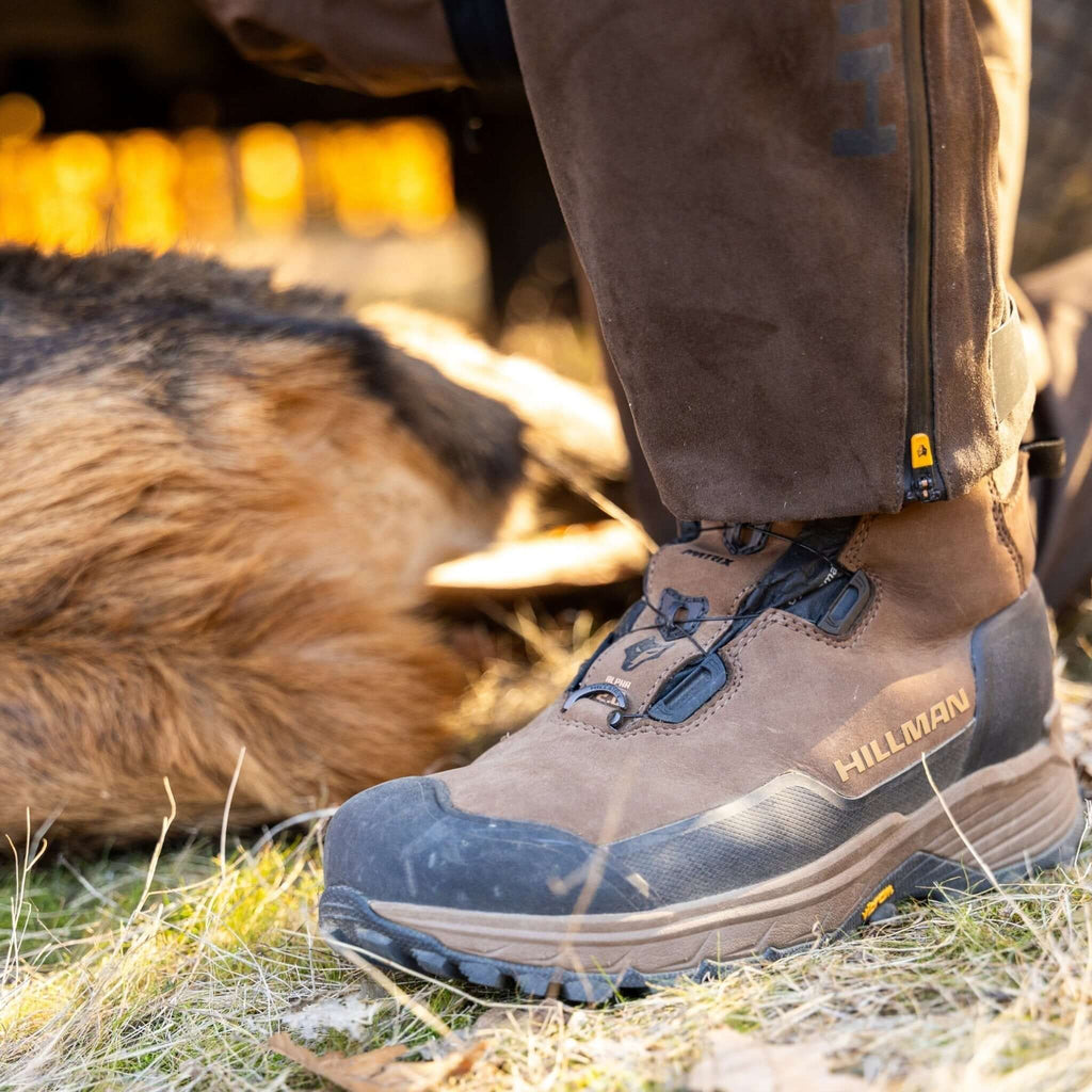 Nahaufnahme einer Person, die Hillman Vorbestellung 2.0 ALPHA DRYHUNT Jagdstiefel trägt - auf Gras, im Hintergrund ein großes Tier, vermutlich ein Hirsch. Lieferung der Stiefel bis 20.07.2025.