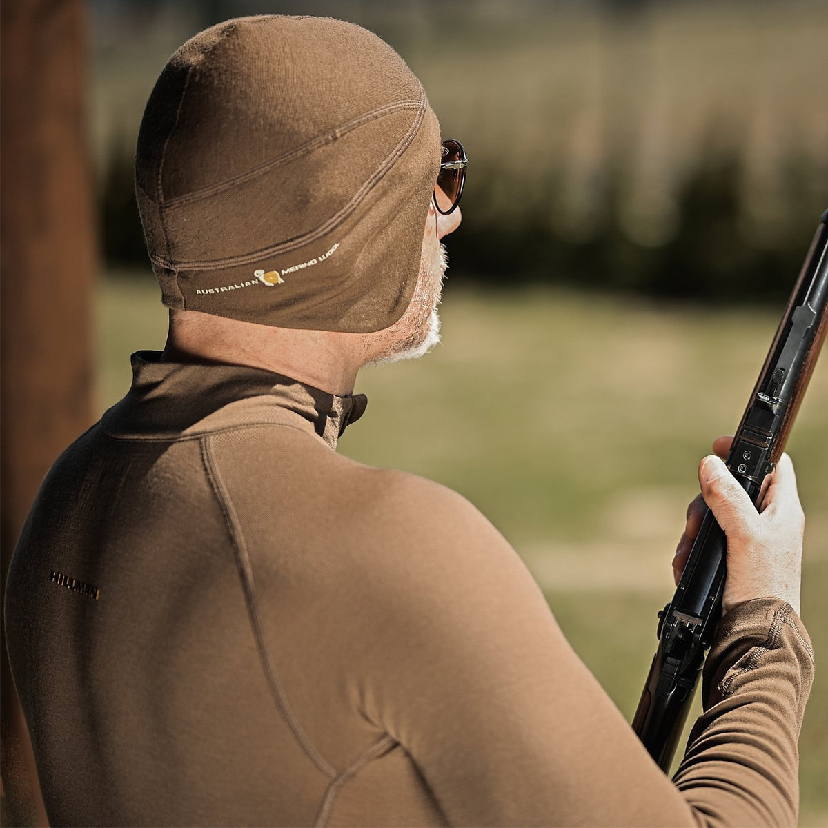 Eine Person in brauner Thermokleidung und einer THERMOMÜTZE zur natürlichen Wärmeregulierung hält im Freien ein Gewehr. Die Sonnenbrille unterstreicht ihren mystischen Auftritt, während sie in eine verschwommene grüne Kulisse blickt. Das Hillman®GWR-Logo ist auf ihrer Ausrüstung präsent, aber dezent.