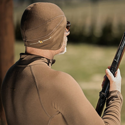 Eine Person in brauner Thermokleidung und einer THERMOMÜTZE zur natürlichen Wärmeregulierung hält im Freien ein Gewehr. Die Sonnenbrille unterstreicht ihren mystischen Auftritt, während sie in eine verschwommene grüne Kulisse blickt. Das Hillman®GWR-Logo ist auf ihrer Ausrüstung präsent, aber dezent.