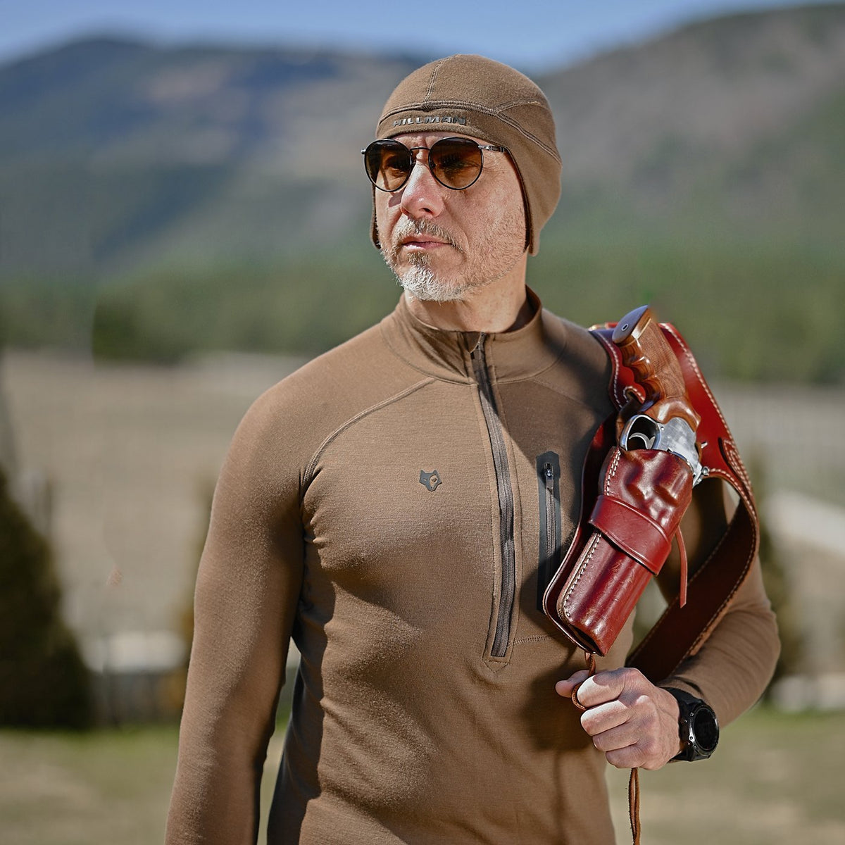 Ein Mann in braunem Outfit und Sonnenbrille, der eine Ledertasche trägt und die Hillman®GWR THERMOMÜTZE – [16μm] Merino Thermo Mütze für Jagd trägt, steht im Freien inmitten majestätischer Berge und Bäume und genießt die natürliche Wärmeregulierung der Natur.