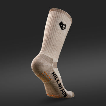 Die JAGDSOCKEN MERINOWOLLE ULTRAWARME [HC] von Hillman®GWR sind beige Socken aus isolierender Naturfaser mit schwarzem Logo auf der Oberseite und dunklem Farbverlauf. Auf der Sohle steht „HILAMAN“ mit orangefarbenen Grip-Punkten und verbindet so wohlige Wärme und Style perfekt.