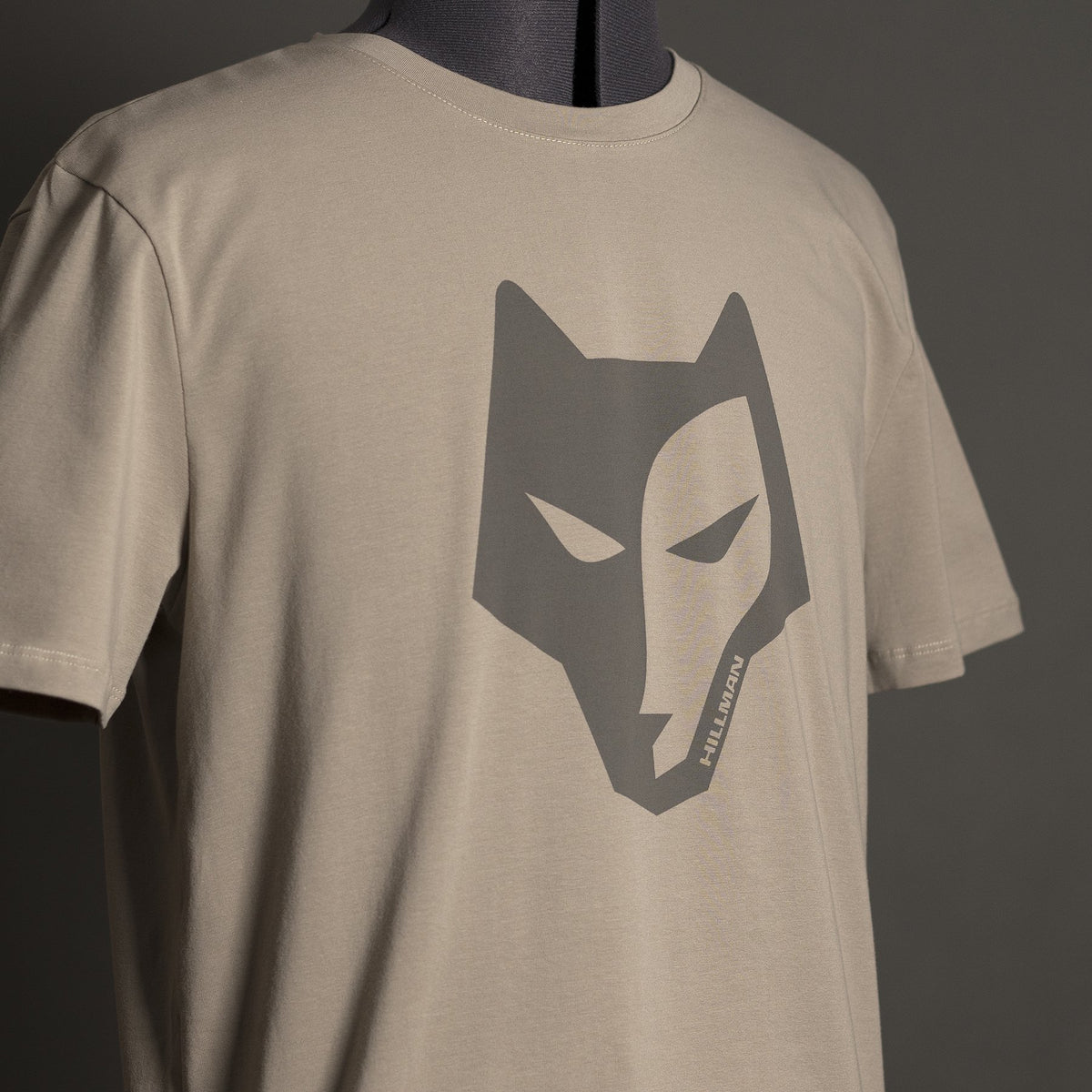 Das Hillman Jagdshirt Alpha - Jagd T-Shirt aus ägyptischer Baumwolle besticht durch eine stilisierte dunkelgraue Wolfsgrafik auf beigefarbenem T-Shirt. Präsentiert auf einer Schaufensterpuppe vor schlichtem dunkelgrünem Hintergrund, vereint es mit seinem kantigen Design Stil und Komfort perfekt.