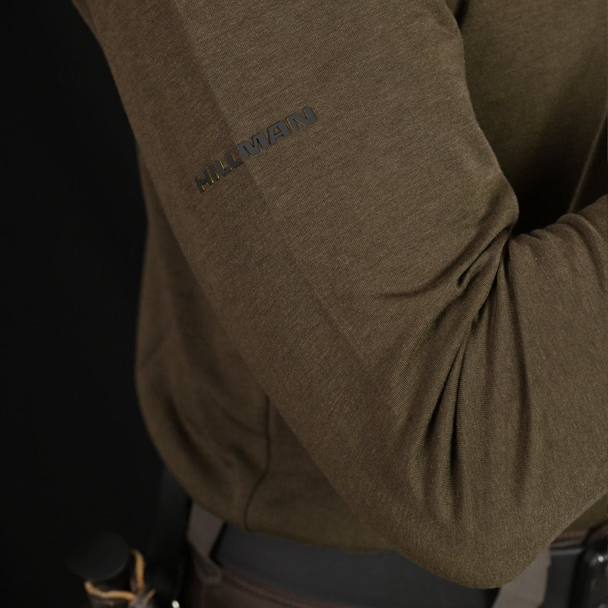 Nahaufnahme einer Person im dunkelgrünen 1UL Langarmshirt Bambus von Hillman. Das antibakterielle und feuchtigkeitsregulierende Material sorgt für hohen Tragekomfort. An ihrer Seite befindet sich ein Ledergürtel mit einem Jagdmesser vor schwarzem Hintergrund.