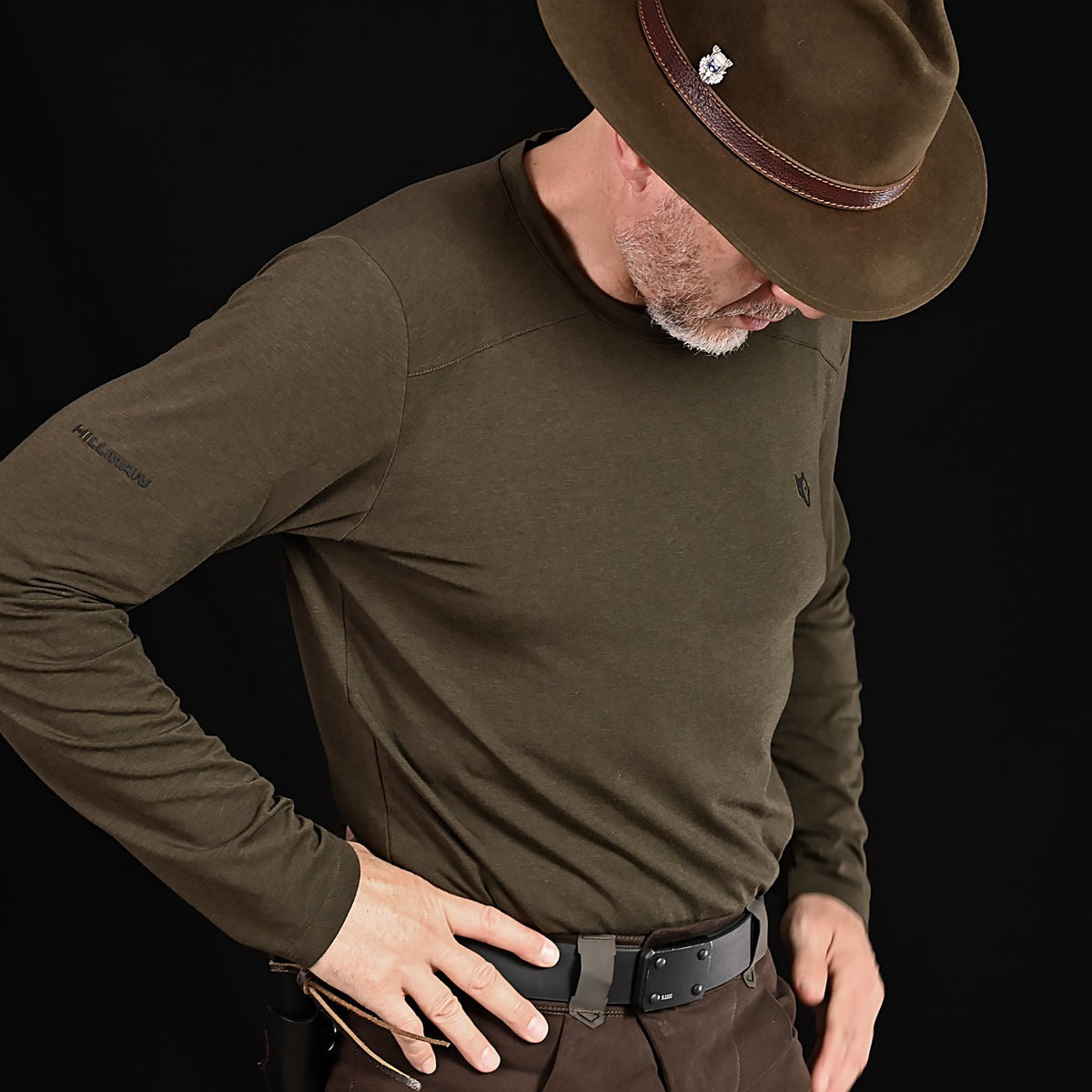 Jemand in einem olivgrünen Hillman 1UL Langarmshirt Bambus, einem breitkrempigen braunen Hut mit silbernem Detail und dunkler Hose rückt vor einem schwarzen Hintergrund einen schwarzen Gürtel zurecht. Sein Gesicht ist verborgen.